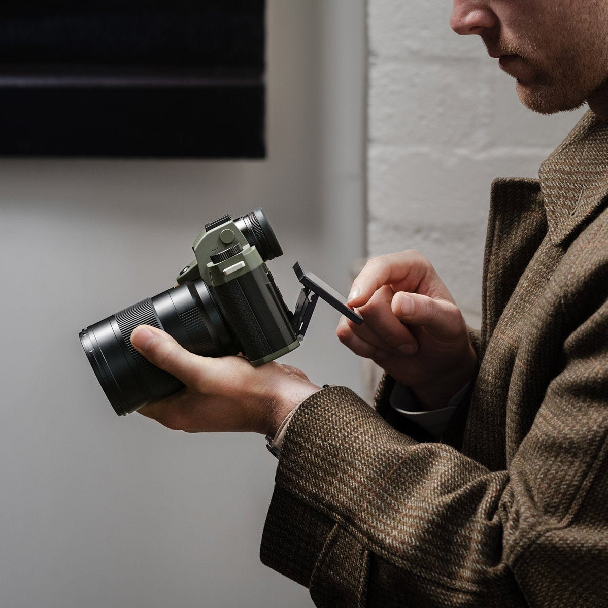Man in a brown coat holding a camera and a small object against a plain background