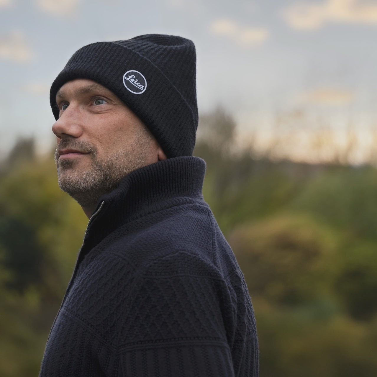 Man wearing a black knit cap with a leica logo, standing outdoors with trees and sky in the background.